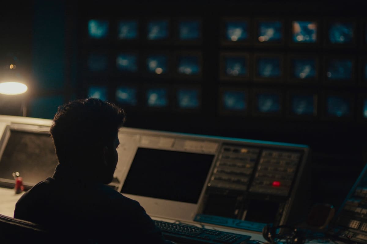A dim news control room, an operator seen from behind facing the monitor wall